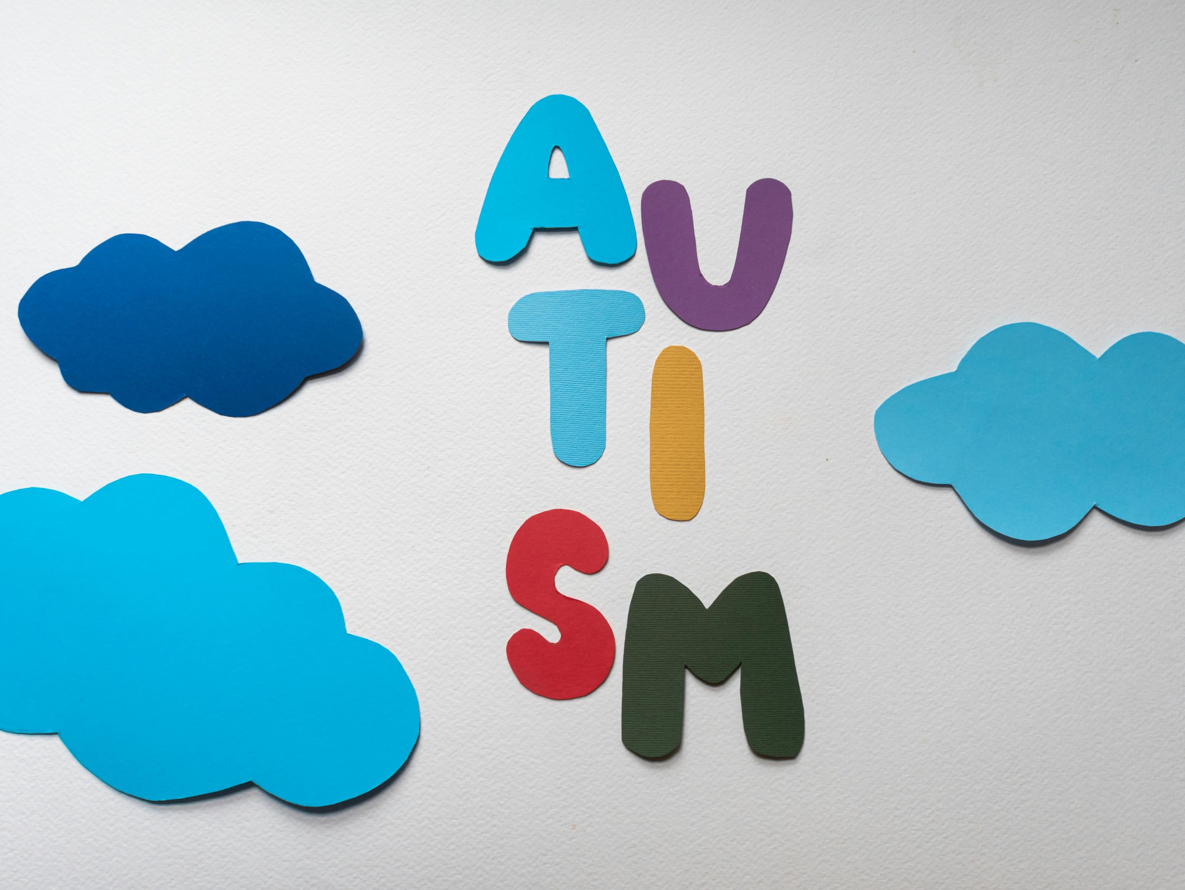 The image shows the word "AUTISM" spelled out vertically with colorful, cut-out letters against a light gray background. The letters are in different colors: "A" in light blue, "U" in purple, "T" in light blue, "I" in yellow, "S" in red, and "M" in dark green. Surrounding the letters are several blue paper cloud cutouts of different shades, arranged decoratively around the word. The composition appears to be a crafted display or educational visual about autism.