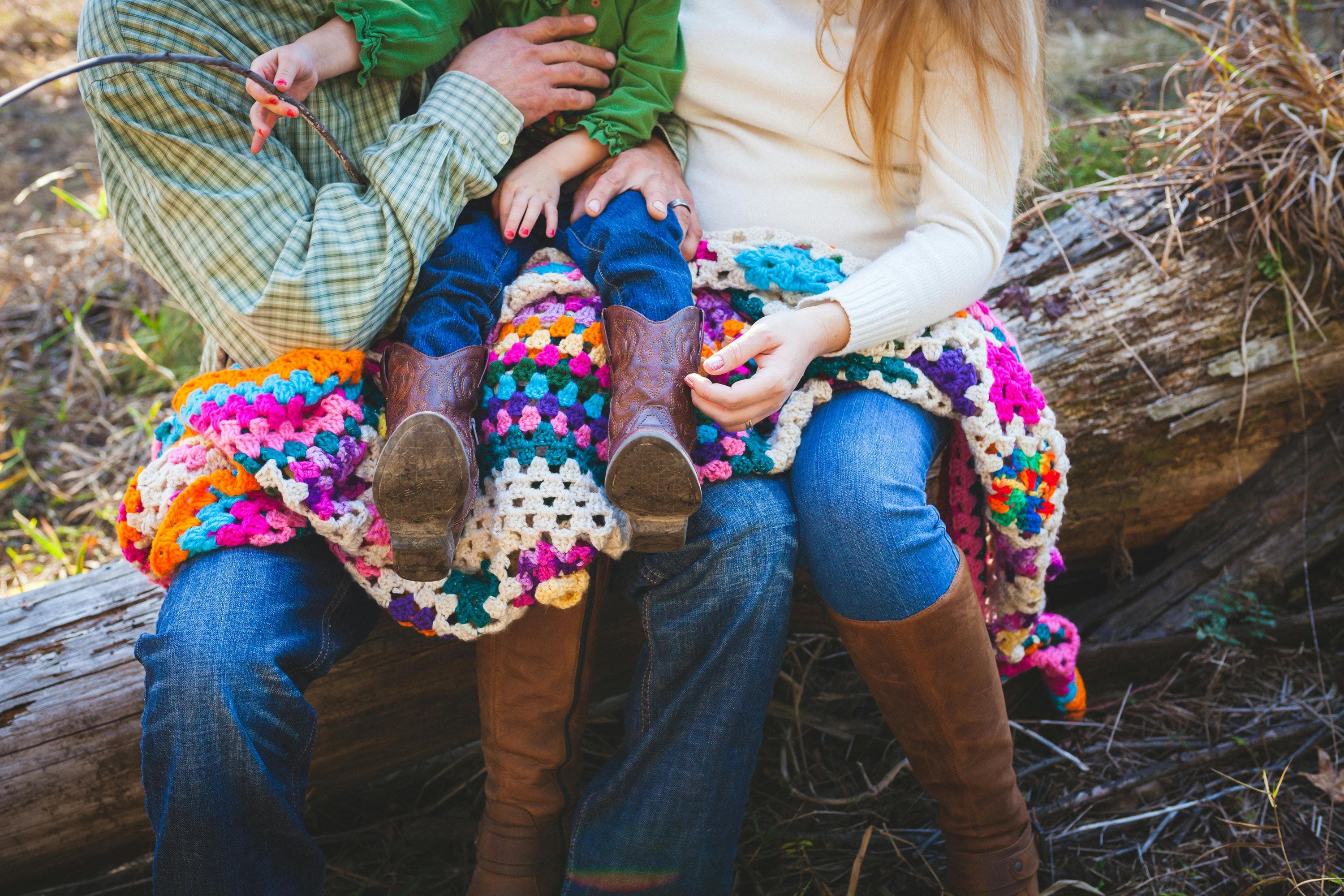 The image shows a family sitting together on a fallen log in what appears to be a wooded outdoor setting. Two adults (only visible from the torso down) are sitting side by side wearing jeans and boots, with a small child seated between them. The child is wearing blue pants and colorful knitted or crocheted leg warmers. All three are partially covered by a vibrant, multicolored crocheted blanket with a granny square pattern featuring bright pinks, blues, yellows, and other colors. The adults' hands are visible holding or supporting the child. The scene suggests a cozy family moment outdoors in autumn or spring.