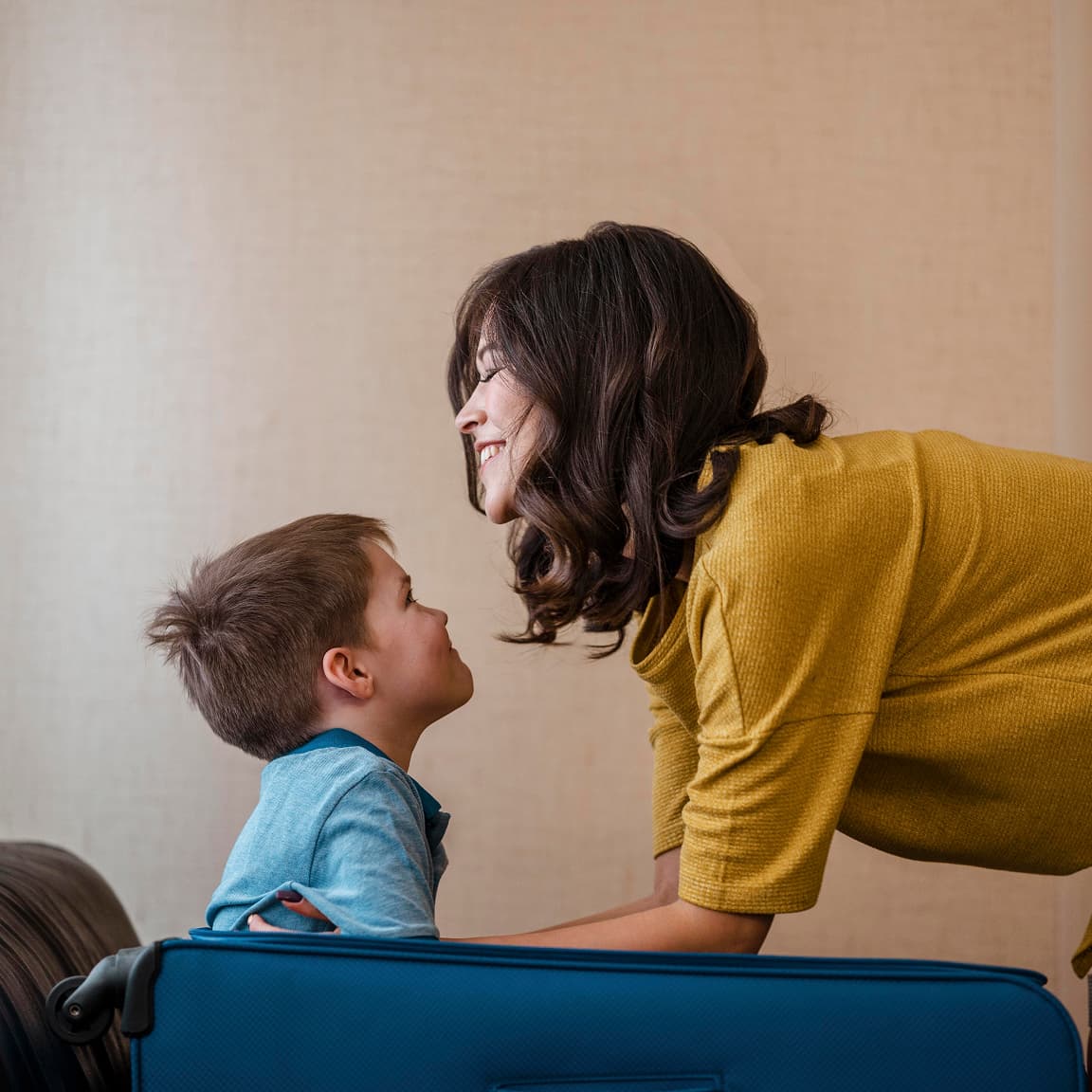A warm and candid moment showing a person in a yellow sweater leaning down to share a joyful interaction with a young child in a light blue shirt, with a blue suitcase visible in the foreground against a neutral background